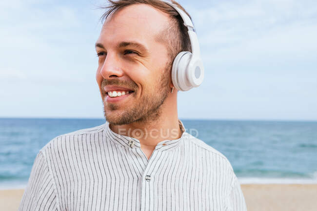 Despreocupado joven barbudo con elegante camisa casual escuchando música a través de auriculares inalámbricos y disfrutando de brisa fresca mientras pasa el día de verano en la playa de arena cerca del mar mirando hacia otro lado - foto de stock