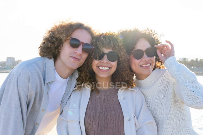 Group of young diverse women and man with curly hair and sunglasses embracing each other while standing on shoreline of cityscape in back lit — Stock Photo
