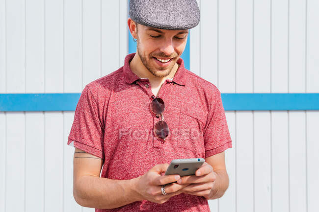 Вміст молодого бородатого хіпстера в повсякденній сорочці поло і кепці перегляду мобільного телефону, стоячи на тлі дерев'яної біло-блакитної стіни на сонячному світлі — стокове фото