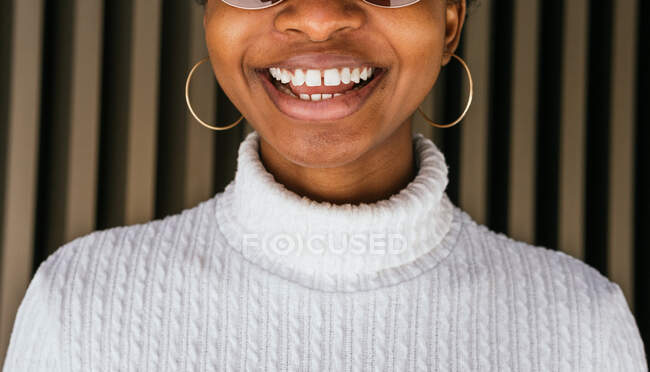 Crop African American female with gapped teeth in trendy sweater smiling happily against striped wall on street — Stock Photo