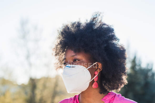 Conteúdo Mulher afro-americana com cabelo encaracolado e respiradora protetora olhando para longe no parque no dia ensolarado durante a pandemia de coronavírus — Fotografia de Stock
