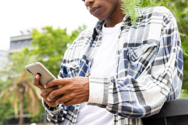 D'en bas copped messages masculins afro-américains méconnaissables sur téléphone portable tout en se tenant dans la rue de la ville — Photo de stock