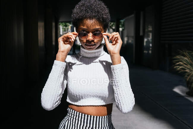 Giovane femmina nera in abito elegante regolazione occhiali da sole e guardando la fotocamera nella giornata di sole sulla strada della città — Foto stock