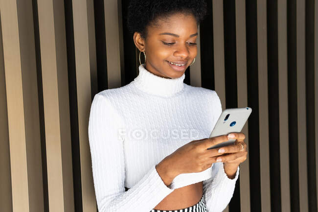 Feliz elegante jovem mulher negra em suéter navegando no smartphone enquanto está perto da parede do edifício listrado na cidade — Fotografia de Stock
