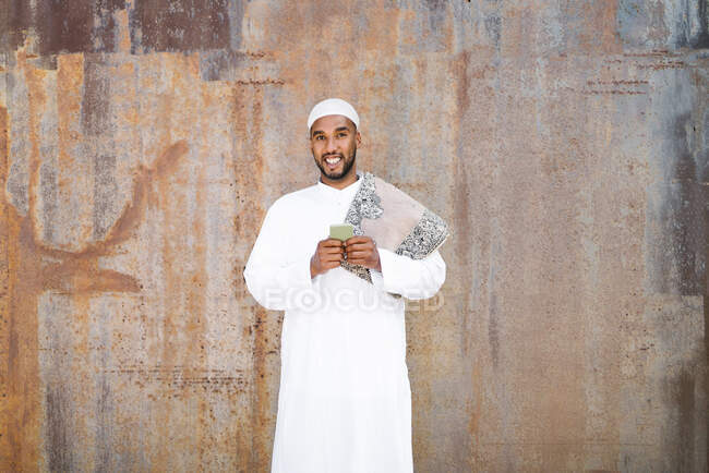 Homme musulman joyeux en vêtements traditionnels souriant et naviguant sur le téléphone portable tout en se tenant près d'un mur minable dans la rue — Photo de stock