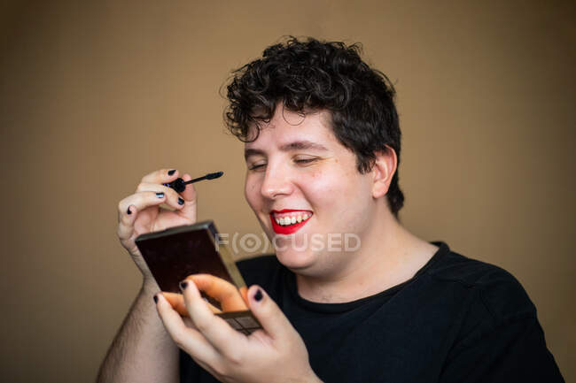 Concentrado excêntrico feminino masculino aplicando rímel com escova ao fazer maquiagem com a boca aberta e segurando espelho — Fotografia de Stock