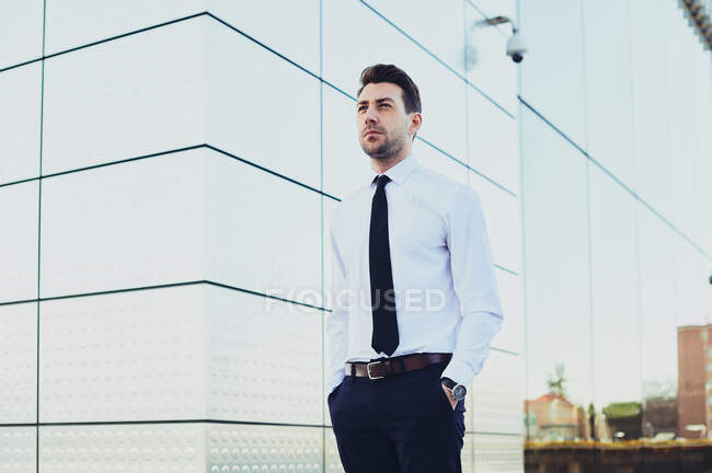 Мужчина-предприниматель в формальной одежде с руками в карманах, смотрящий в сторону в городе — стоковое фото