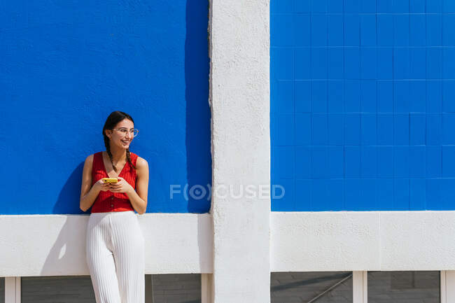 Контент стильна жінка спирається на яскраво-блакитну стіну на вулиці і переглядає соціальні медіа на мобільний телефон в сонячний день влітку — стокове фото