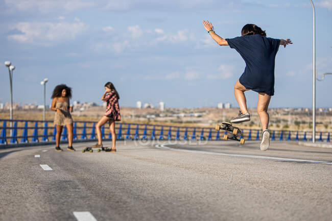 Unrecognizable young woman doing a trick with her long board by a bridge with her companions in the background — Stock Photo