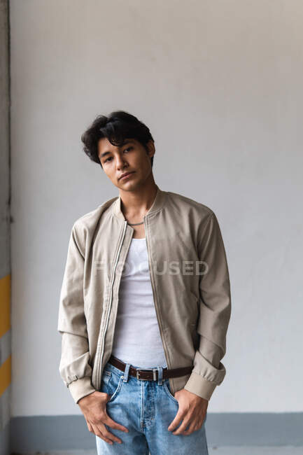 Retrato de un joven latino con ropa casual mirando con confianza a la cámara con las manos en los bolsillos mientras está parado en el estacionamiento - foto de stock