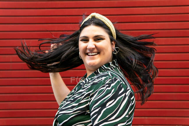 Side view of cheerful young curvy brunette in stylish striped outfit and headband shaking long hair and looking at camera against red wall — Stock Photo