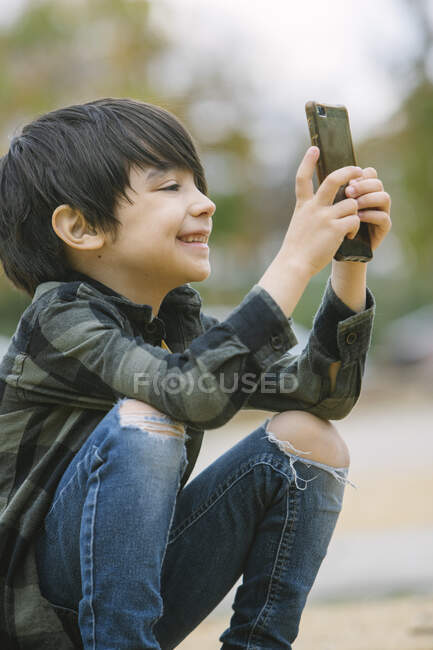 Side view of adorable boy in casual clothes browsing cellphone while sitting on skateboard on pedestrian crossing in city — Stock Photo