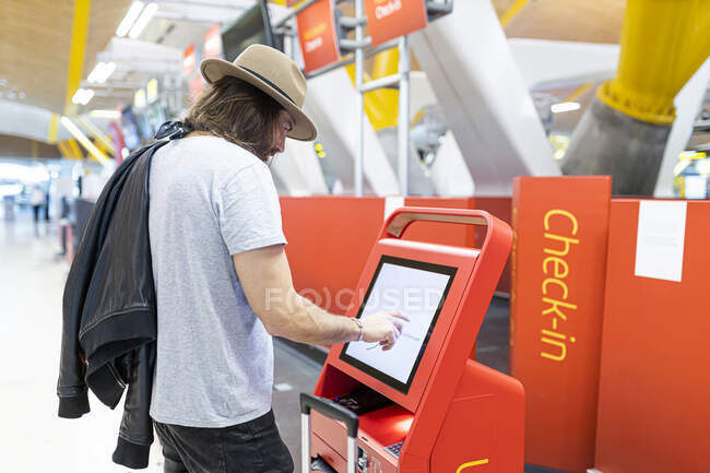 Young man with beard and long hair and hat buying airline tickets at the airline's automatic terminal — Stock Photo