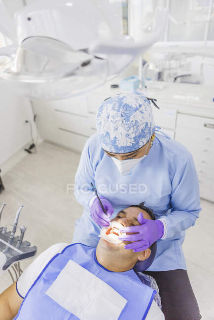 Dall'alto di dottore femminile anonimo in denti di pulizia uniformi di paziente maschile con riavvolgitore dentale in ospedale — Foto stock