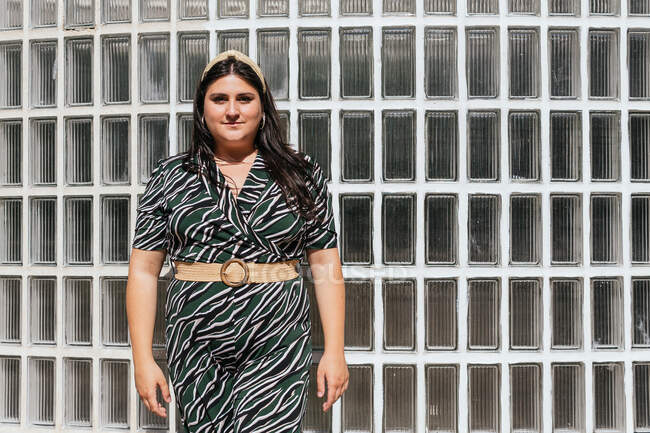 Young curvy female model in stylish black dress with diagonal striped print and belt standing against glass wall on street — Stock Photo