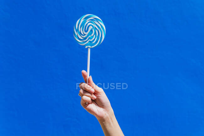 Crop hand of unrecognizable female with delicious lollipop on stick against blue wall in city on sunny day — Stock Photo