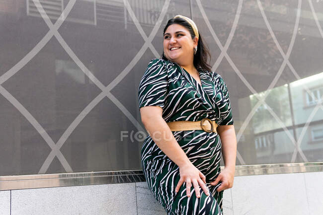Happy young curvy female in stylish ornamental dress and laughing while standing against wall of modern building — Stock Photo