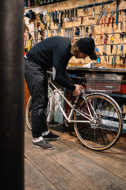 Vista lateral da bicicleta de fixação mestre masculino ocupado enquanto trabalhava na oficina de reparo pobre — Fotografia de Stock