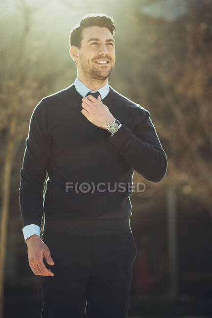 Hombre empresario barbudo sonriente en reloj de pulsera con corte de pelo moderno mirando hacia otro lado en la ciudad en la parte posterior iluminada - foto de stock