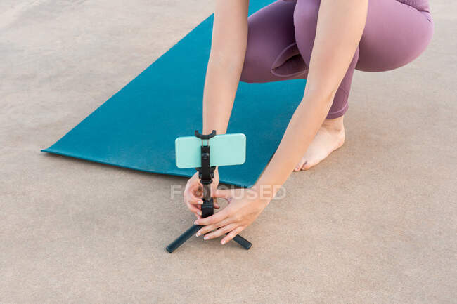Angle élevé de la femelle méconnaissable recadrée préparant téléphone portable sur trépied pour faire du yoga pendant la leçon en ligne — Photo de stock