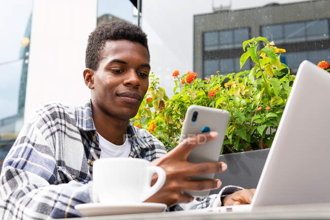 Low angle of content African American male freelancer browsing smartphone and working remotely in outdoors cafe while sitting at table with cup of coffee and laptop — Stock Photo