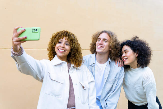 Group of young diverse women and man with curly hair using phone to take selfie together against beige wall — Stock Photo