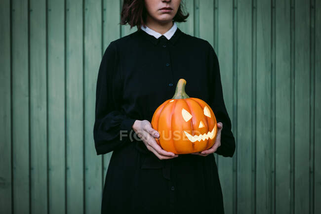 Обрезанный неузнаваемая женщина в черном платье и с Хэллоуин тыквы фонарь стоит напротив деревянной стены дома — стоковое фото