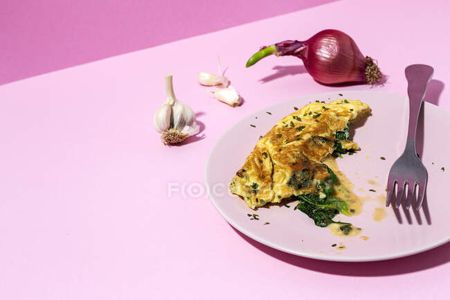 Omelete saborosa na placa contra raminhos de salsa frescos e cebola vermelha com dentes de alho no fundo rosa — Fotografia de Stock
