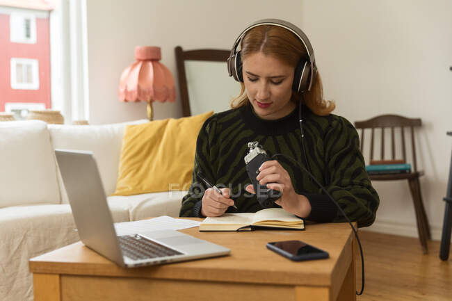 Host radio femminile focalizzato con microfono e cuffie che scrivono nel blocco note mentre si preparano per la registrazione del podcast a casa — Foto stock