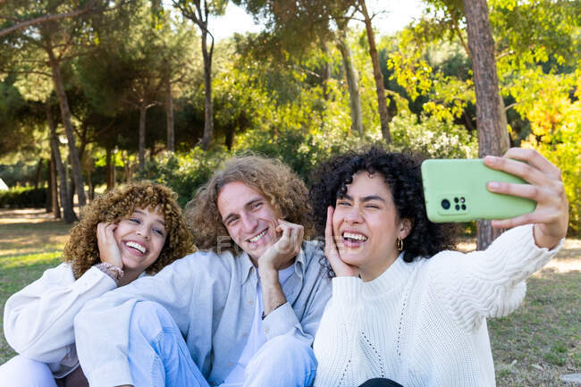 Grupo de mujeres multirraciales jóvenes felices y hombre con el pelo rizado sentado en la hierba verde en el parque mientras toma selfie con el teléfono móvil - foto de stock