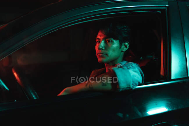 Young latin man sitting in car and looking at camera from driving seat under color lights at night — Stock Photo