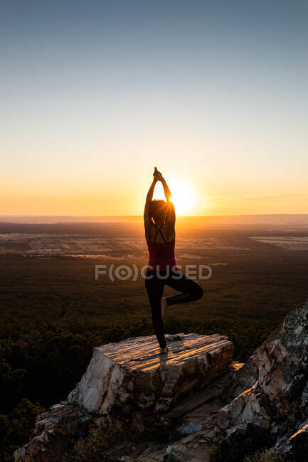 Joven yogi practicando yoga sobre una roca en la montaña con la luz del amanecer, vista trasera con una pierna y brazos levantados - foto de stock