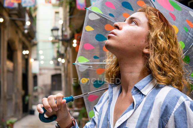 Dal basso curioso maschio elegante con i capelli lunghi in piedi sotto l'ombrello trasparente in strada il giorno di pioggia e guardando altrove — Foto stock