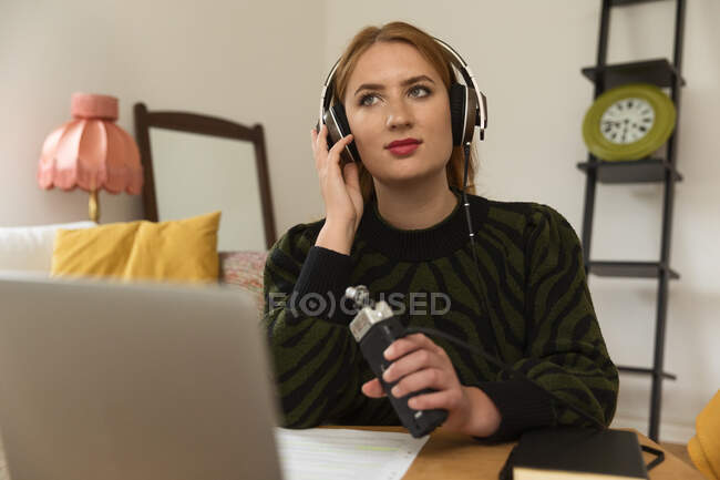 Задумчивая женщина в наушниках записывает подкаст, разговаривая в микрофоне и используя ноутбук дома, отворачиваясь — стоковое фото