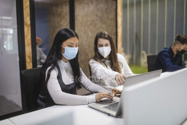 Multirassische Kolleginnen mit Masken sitzen mit Laptop am Tisch und diskutieren Geschäftsprojekte, während sie im Coworking Space arbeiten — Stockfoto