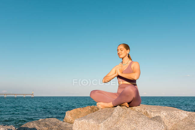 Mujer serena con los ojos cerrados haciendo yoga en pose de loto durante la meditación sobre roca en la orilla del mar - foto de stock