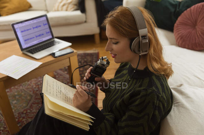Боковой вид сфокусированной женщины-радиоведущей с микрофоном и наушниками, записывающей в блокнот во время подготовки к записи подкаста дома — стоковое фото