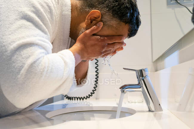 Vista lateral da colheita macho de meia idade em roupão de banho branco limpeza rosto com água ao fazer rotina matinal no banheiro — Fotografia de Stock