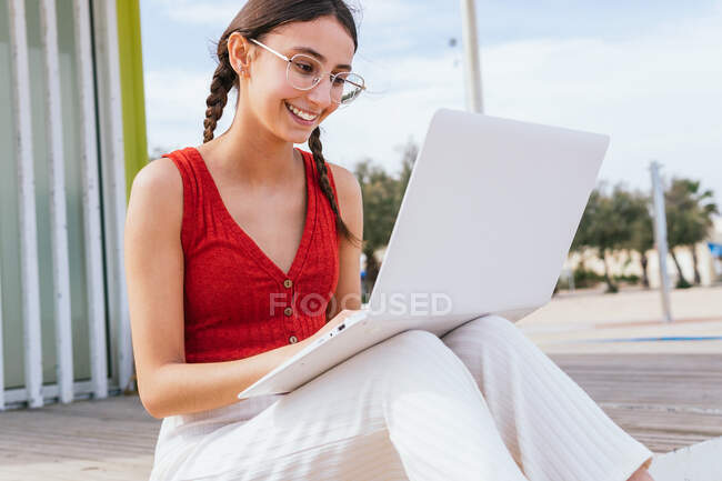 Contenido freelancer femenina sentada en la orilla del mar y escribiendo en el portátil mientras trabaja remotamente en el proyecto en verano - foto de stock