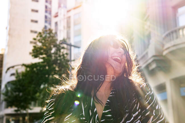 Niedrige Winkel der fröhlichen jungen Frau in stilvollem Outfit und Ohrringe schütteln Haare im Sonnenlicht auf der städtischen Straße — Stockfoto