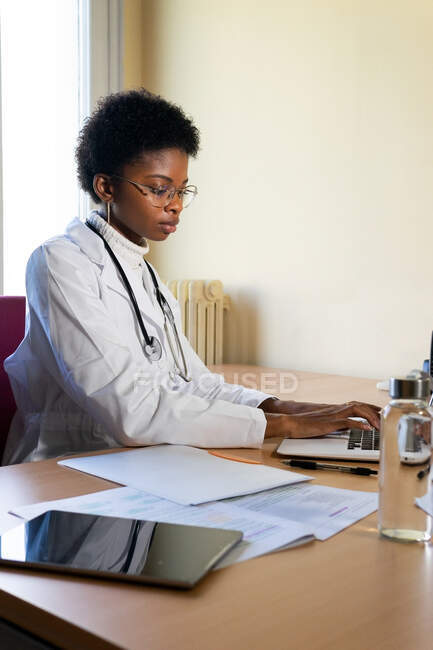 Joven médico afroamericano escribiendo informe sobre el ordenador portátil mientras trabaja en la mesa en la oficina clínica moderna - foto de stock