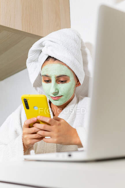 Giovane femmina con maschera di argilla seduta a tavola con computer portatile e navigando sui social media sul cellulare durante la sessione spa a casa — Foto stock