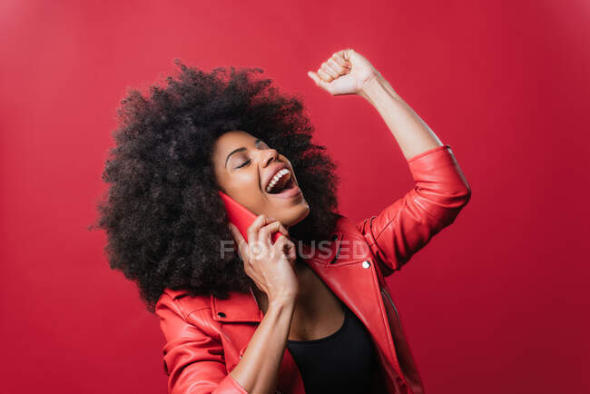 Eccitato donna afroamericana con pugno serrato fare chiamata sul telefono cellulare e celebrare il successo su sfondo rosso — Foto stock