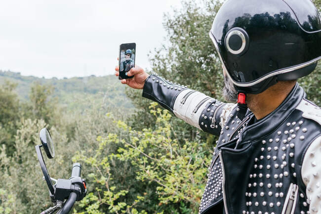 Side view of unrecognizable bearded male biker in trendy leather jacket with rivets and protective helmet taking self portrait while sitting on motorcycle parked near lush green forest — Stock Photo