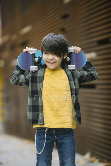 Cheerful boy in casual clothes smiling and looking away while standing with skateboard behind head on street against blurred background — Stock Photo