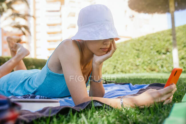 Vista lateral da jovem usando o telefone enquanto estava deitado na grama em um dia de verão — Fotografia de Stock
