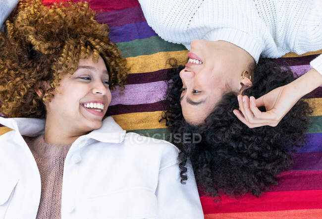 De cima de namoradas multirraciais felizes com cabelo encaracolado deitado em xadrez colorido no parque e olhando um para o outro enquanto descansam juntos — Fotografia de Stock