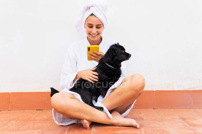 Glückliche Hündin in Bademantel und Handtuch streichelt schwarzen Hund und surft Handy, während sie im Schneidersitz auf dem Boden während der Hautpflege-Routine sitzt — Stockfoto