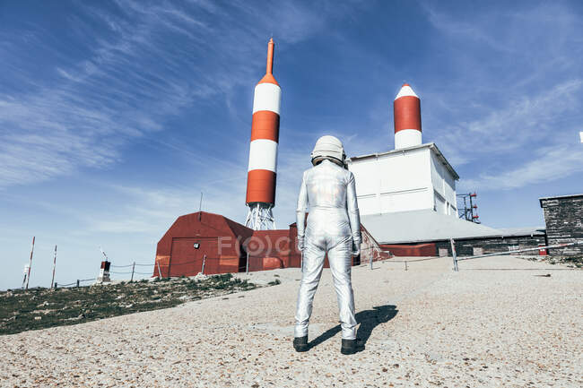 Back view man in spacesuit standing on rocky ground against metal fence and striped rocket shaped antennas on sunny day — Stock Photo