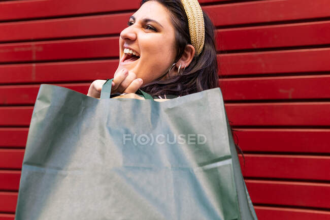 Side view of happy young curvy female carrying shopping bags while standing near red striped wall in city — Stock Photo